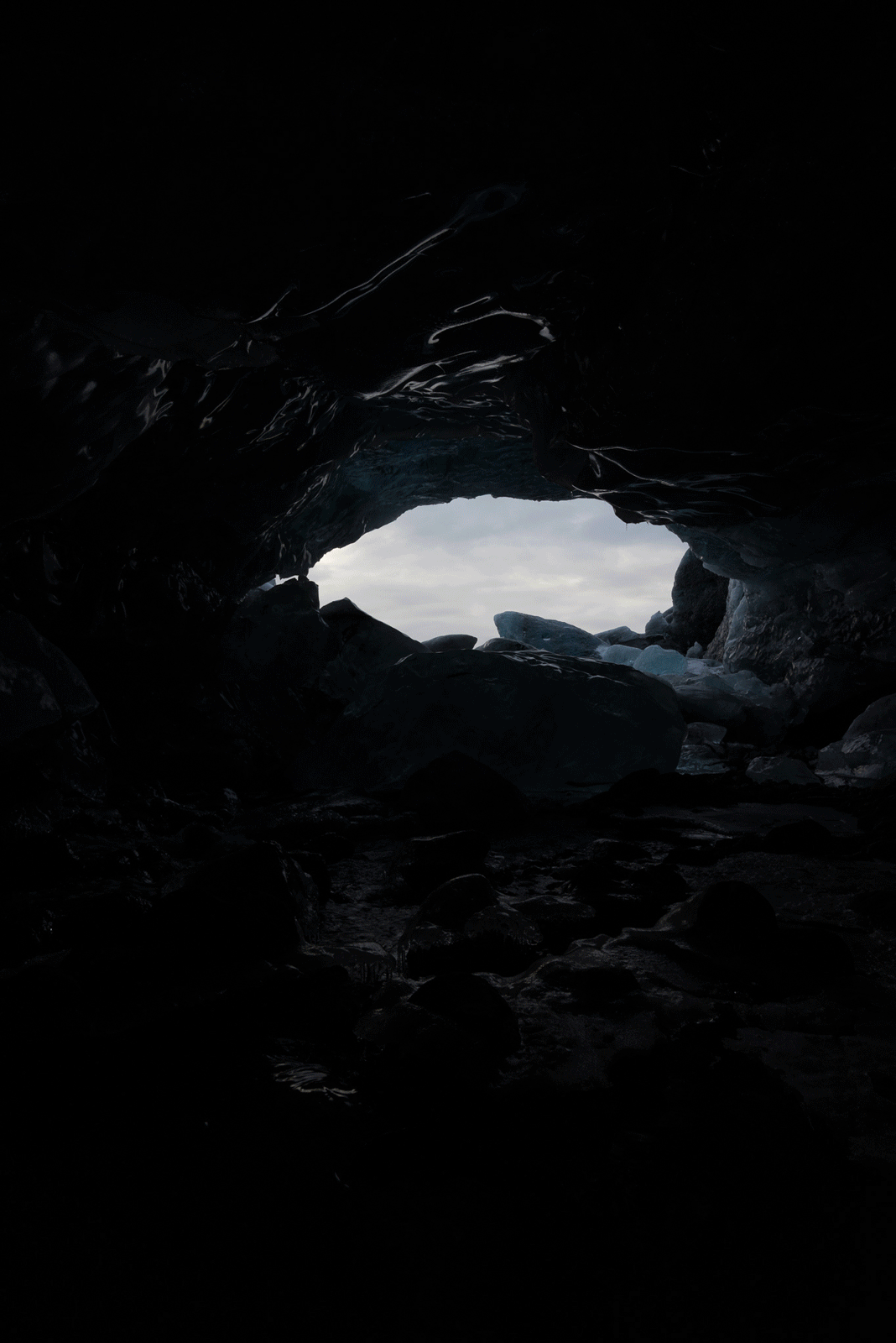 View from inside a dark cave looking out toward a bright, cloudy sky. The cave entrance is irregularly shaped, and rocks and uneven ground are visible in the shadows at the cave’s mouth.