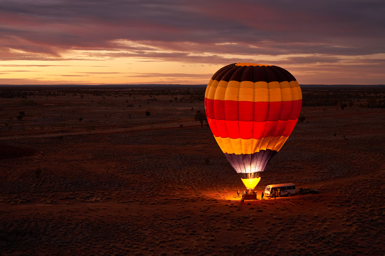 A colorful hot air balloon is being inflated near a white van on a vast, open plain at sunrise or sunset, casting a warm glow on the landscape under a dramatic sky.