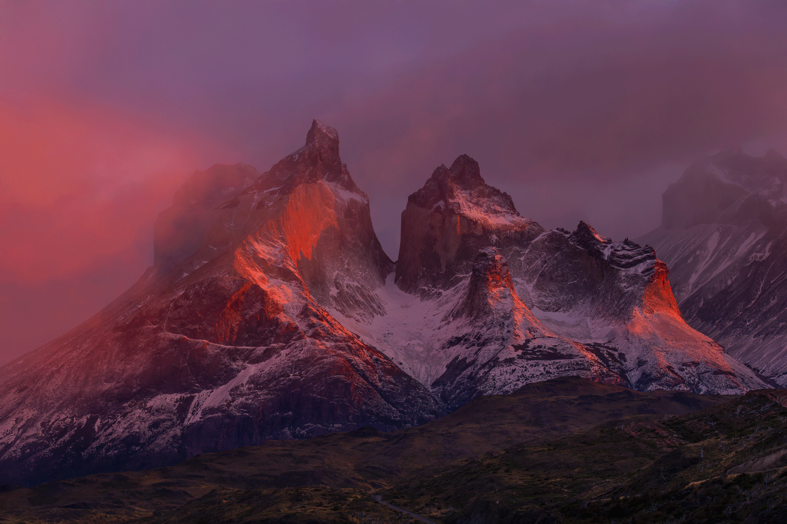 Dramatic snow-capped mountain peaks are bathed in warm red-orange light at sunrise or sunset, under a moody, cloudy sky. The rugged landscape below is shadowed with hints of green.