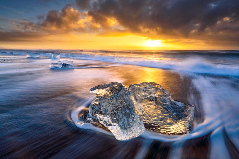 Clear ice chunks rest on a black sand beach at sunrise, with golden sunlight reflecting off the wet sand and waves, and dramatic clouds overhead.