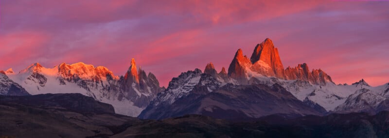 Snow-capped mountains glow orange and pink under a dramatic sunrise sky with streaks of purple and red clouds above rugged peaks and ridges.