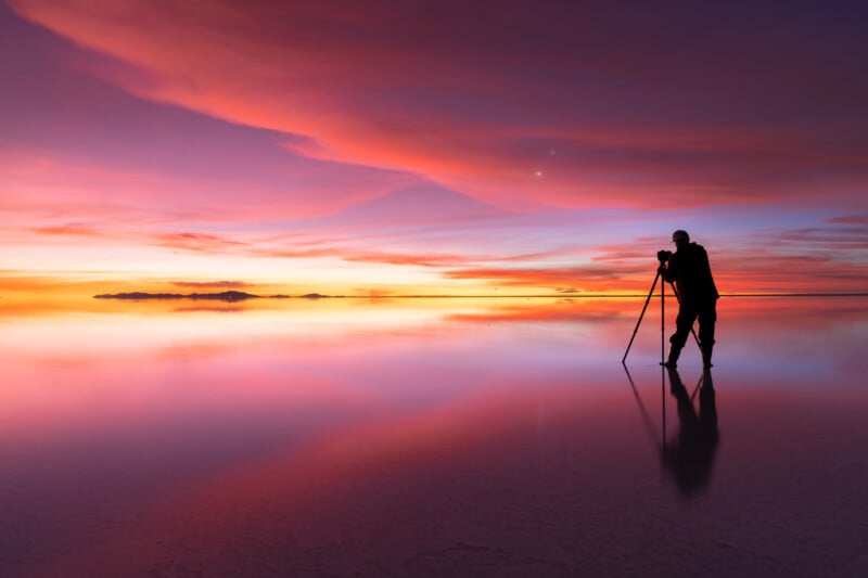 A person with a camera on a tripod stands on a reflective, colorful surface at sunset, surrounded by vibrant pink, orange, and purple clouds.