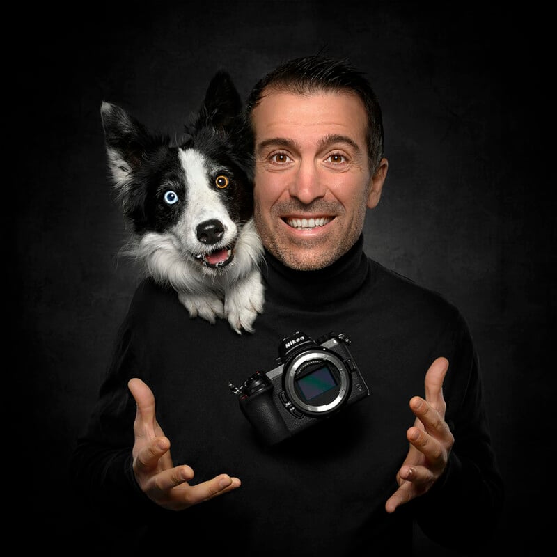 A smiling man in a black turtleneck poses with a black and white dog resting its head on his shoulder. The man holds a camera floating between his hands, and the dog has one blue and one brown eye.