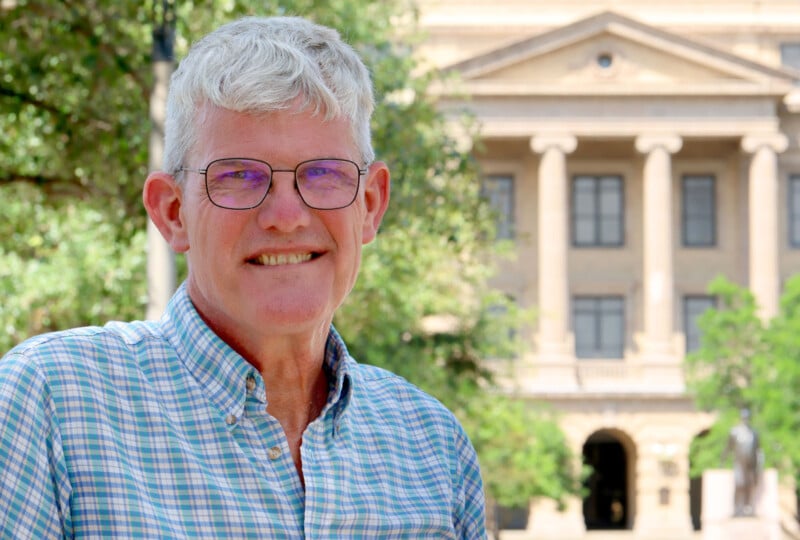 A smiling older man with gray hair and glasses stands outdoors in front of a historic building with columns, wearing a plaid shirt. Trees and greenery are visible in the background.