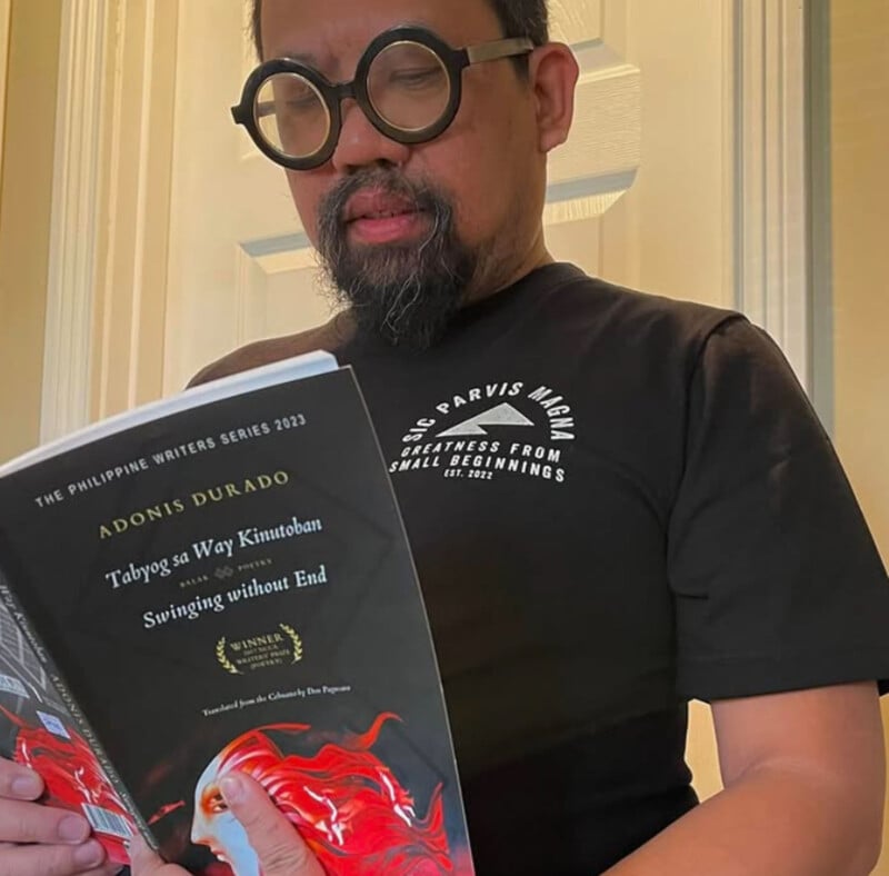 A man with a beard and round black glasses is reading a book titled "Taboya sa Way Kinutuban or Swinging without End" by Adonis Durado. He is wearing a black t-shirt and standing indoors.