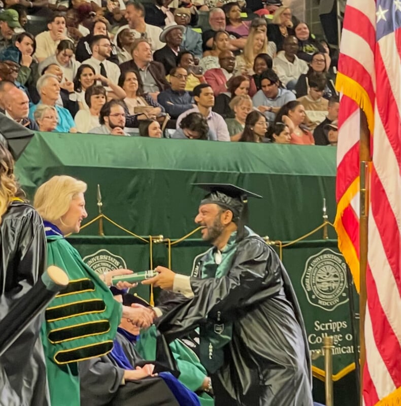 A graduate in a cap and gown shakes hands with a faculty member while receiving a diploma at a graduation ceremony. The audience is seated in the background. An American flag and Scripps College banners are visible.