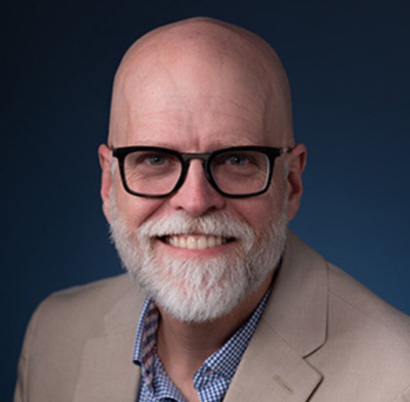 A smiling older man with a bald head, white beard, and black glasses, wearing a beige blazer over a blue checkered shirt, posed against a dark blue background.