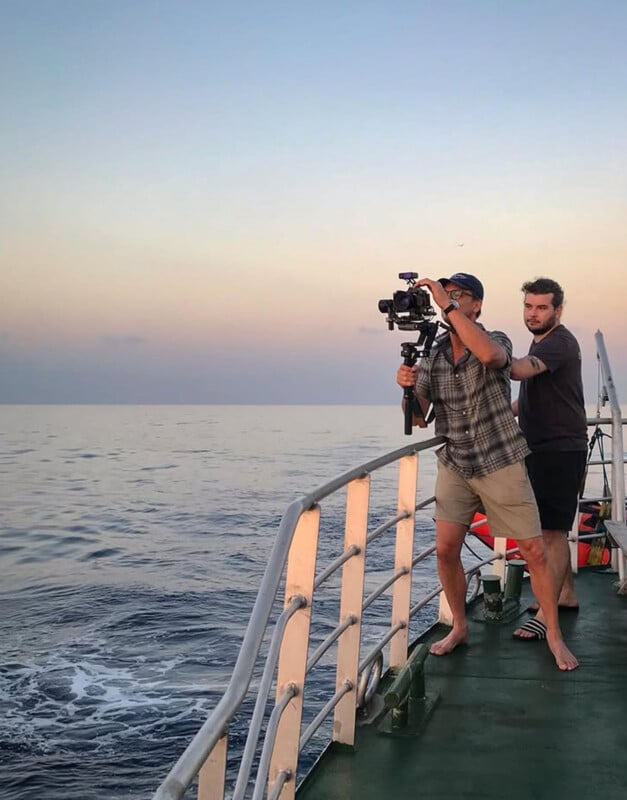 Two people stand barefoot on a boat deck at sunset, one operating a camera on a stabilizer, filming over the ocean while the other holds onto him for support. The sky is clear with soft pastel colors.
