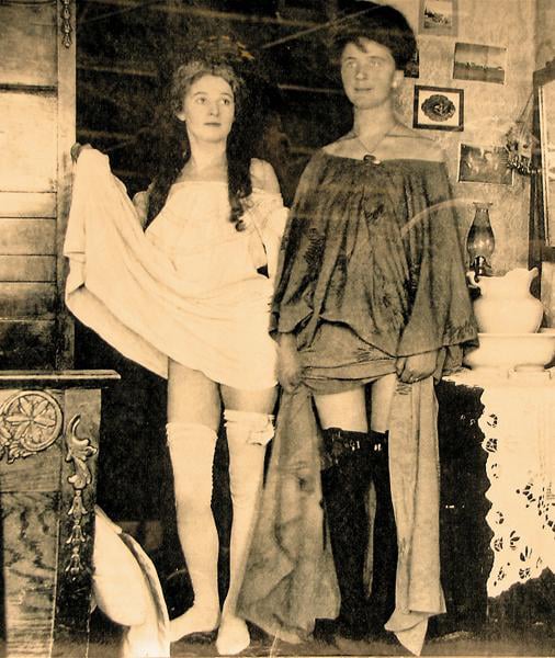 Two women in vintage attire and stockings pose indoors, holding up their skirts. The sepia-toned photo shows a cluttered room with shelves, framed pictures, and various household items in the background.