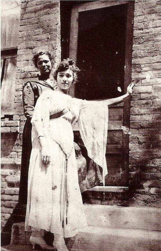 Two women, one standing behind the other, pose in front of a brick building. The woman in front wears a light-colored dress and leans with one arm on a doorframe; the woman behind wears a dark dress and stands on the step.