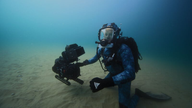 A diver in a blue wetsuit and full-face mask kneels on the sandy ocean floor while holding an underwater camera rig, surrounded by blue water and low visibility.