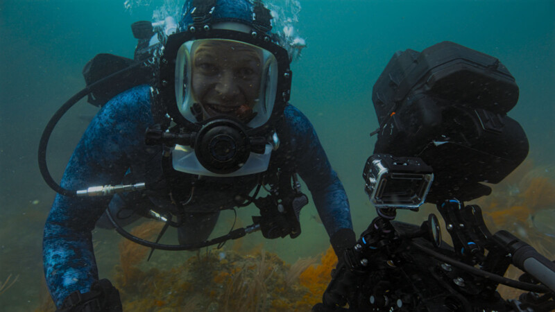 A scuba diver in a blue wetsuit and full-face mask smiles underwater while looking at a camera rig on the ocean floor, surrounded by aquatic plants and blue-green water.