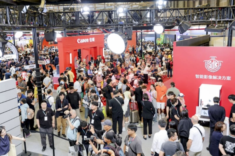 A large crowd attends a busy technology or camera expo with numerous booths, including a prominent red Canon booth, bright lights overhead, and people interacting or exploring displays.