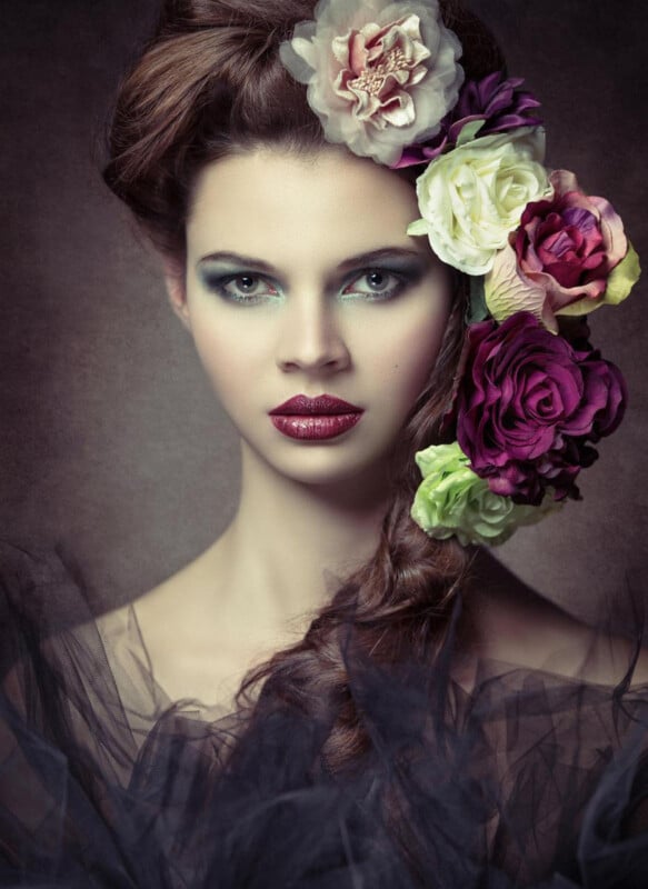 A woman with striking makeup and piercing eyes wears her hair in an elaborate style adorned with large, colorful roses and flowers. She poses against a dark, textured background, with sheer black fabric draped around her shoulders.