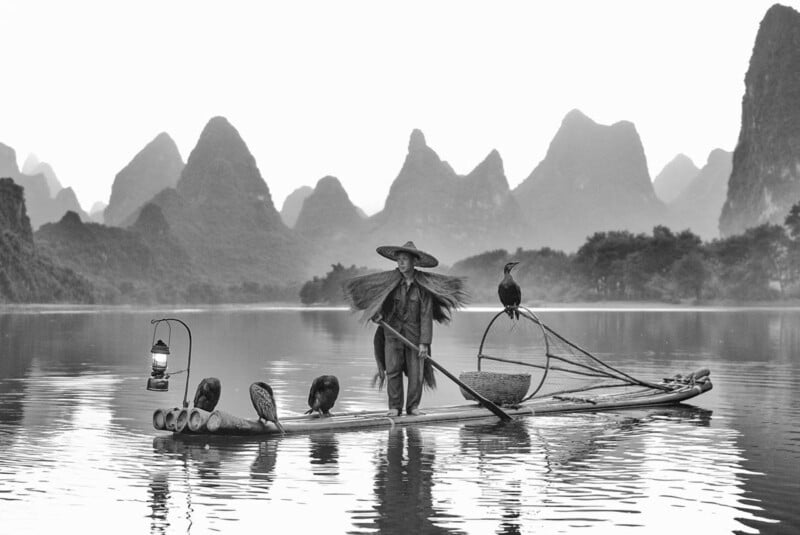 A fisherman wearing traditional attire stands on a bamboo raft with cormorant birds on a calm river, surrounded by karst mountains and mist, with a lantern hanging from the raft.