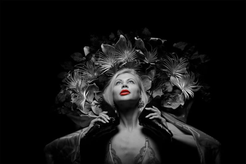 A woman in black and white poses dramatically with red lips emphasized. She looks upward, framed by an elaborate headdress made of large metallic flowers and leaves against a dark background.