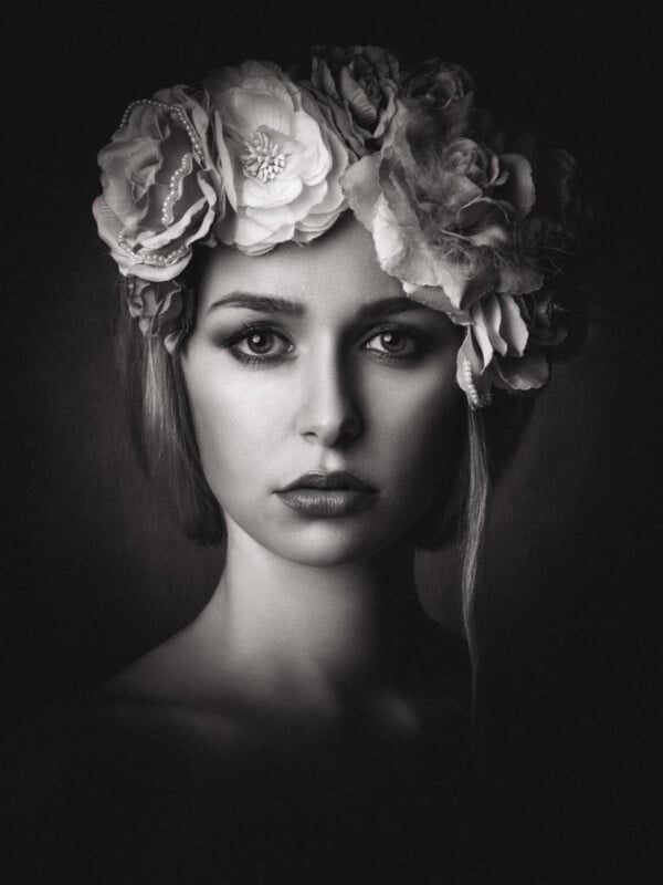 Black and white portrait of a woman with intense gaze, wearing a large floral crown. The lighting highlights her facial features while the background fades to darkness.