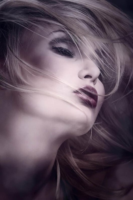 A close-up portrait of a woman with dramatic makeup, dark lipstick, and eyeshadow. Her blonde hair sweeps across her face, partially obscuring her closed eyes, creating a soft, artistic, and ethereal effect.