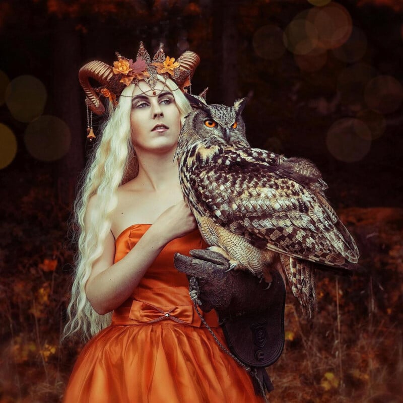 A woman in an orange dress and a horned, autumn-themed crown stands in a forest holding a large owl on her gloved arm. Her long, wavy blond hair contrasts with the dark, blurred trees in the background.