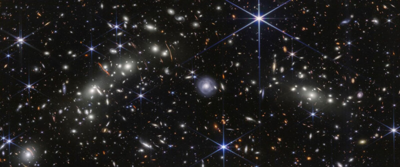 A deep space image showing countless distant galaxies of various shapes and sizes, scattered against a dark background with several bright stars featuring diffraction spikes.