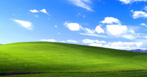 A bright green grassy hill under a blue sky with scattered white clouds. The scene is sunlit and peaceful, with no trees or buildings visible.