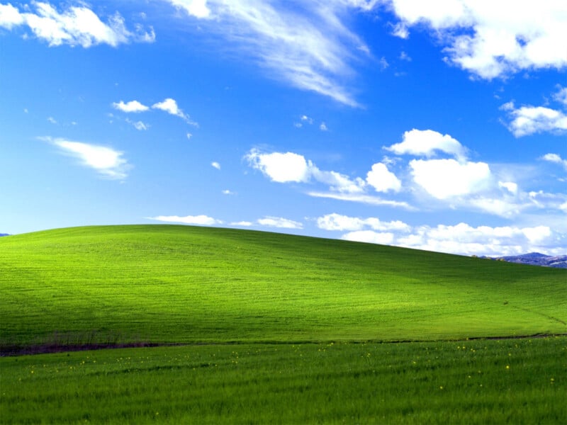 A green grassy hill under a bright blue sky with scattered white clouds, reminiscent of a classic computer desktop wallpaper.