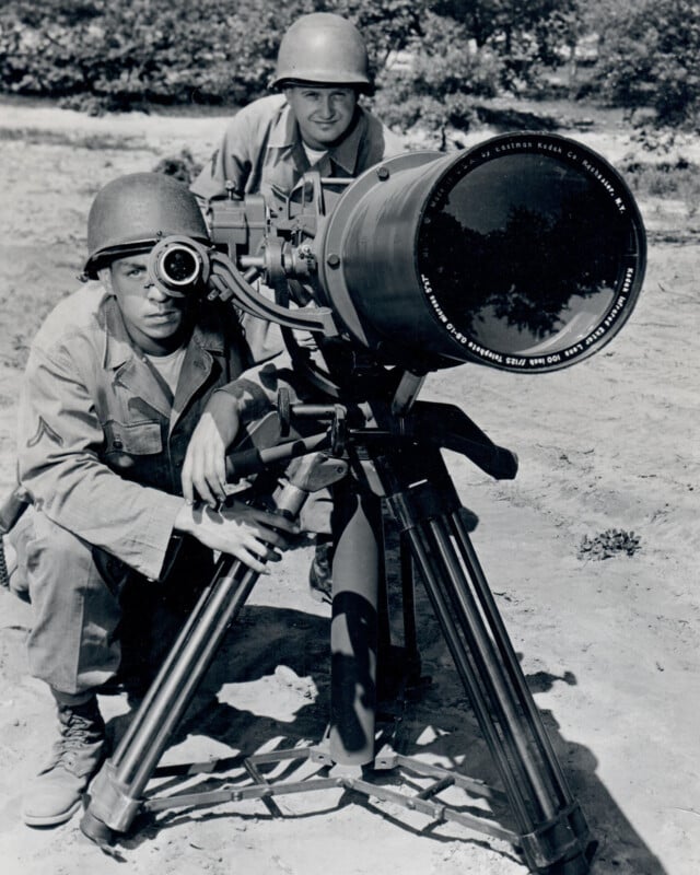 Two soldiers in military uniforms and helmets operate a large camera on a tripod outdoors. One soldier kneels and looks through the viewfinder, while the other stands behind, observing. They appear focused and prepared in a natural setting.