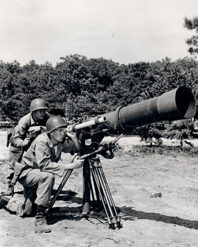 Two soldiers in military uniforms and helmets operate a large mounted telescope or optical device outdoors, with trees in the background. The front soldier adjusts the lens, while the second soldier watches closely.