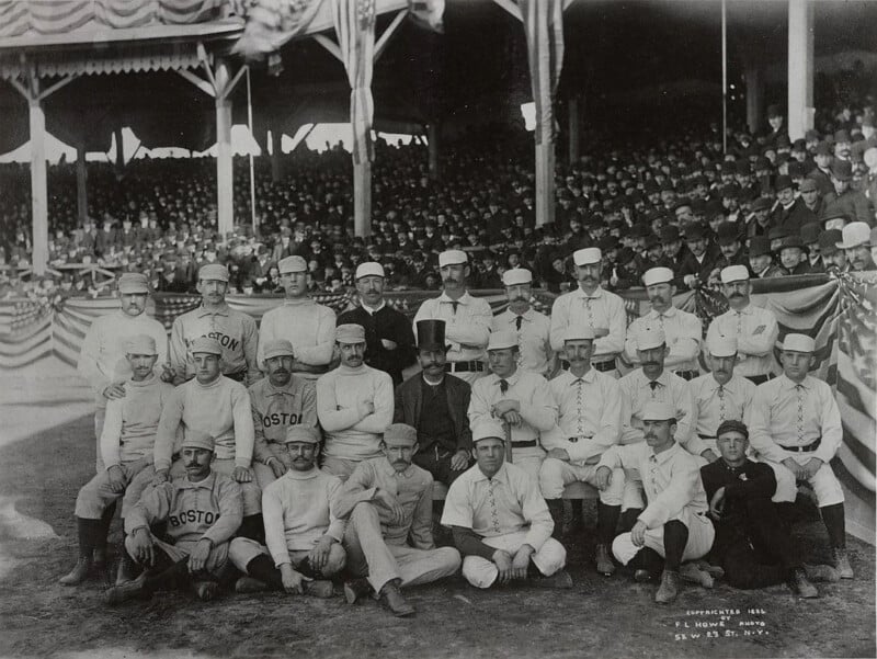 A black and white photograph shows a group of baseball players in uniforms, positioned in front of a crowded stadium. Some individuals are seated while others stand. The players wear uniforms with "Boston" printed on their shirts. Spectators fill the stands in the background.