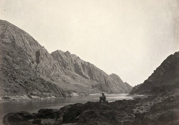 Man sitting on a rocky shore beside the Colorado River in Iceberg Canyon, on the border of Mojave County, Arizona and Clark County, Nevada. Taken in 1871.