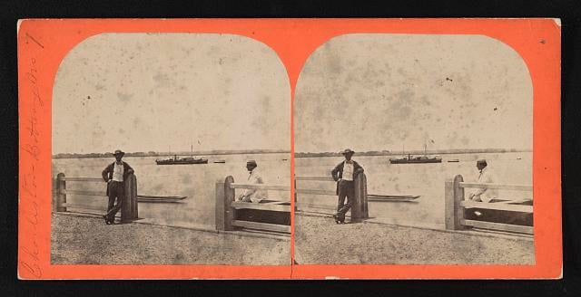 Photograph shows a view of Charleston harbor from the battery with a ship at anchor in the distance. One man leans against the fence post while another is seated on a bench.