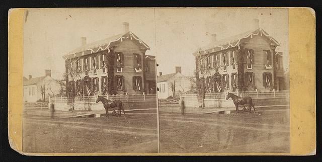 Photograph shows a street view Abraham Lincoln's home in Springfield, Illinois draped in mourning on the day of his funeral. A horse stands near a wooden plank from the street to the sidewalk.