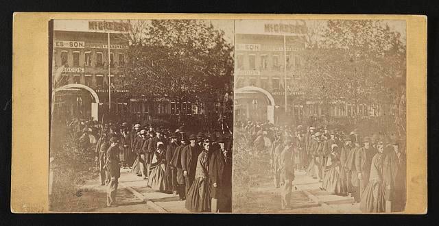 Photograph shows a group of citizens entering the grounds of the Illinois state house to view the body of Abraham Lincoln on May 3 or 4, 1865. Two soldiers stand near the specially built arch. The African American man with the cane near the head of the line is Reverend Henry Brown.