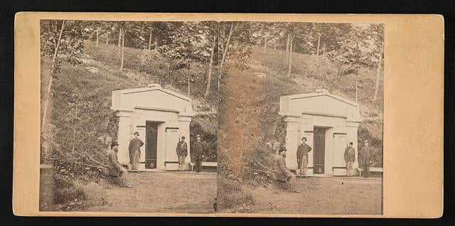 Photograph shows the public receiving tomb of Abraham Lincoln at Oak Ridge Cemetery. One man is seated in the foreground and another man stands at the entrance to the vault along with two soldiers in uniform.