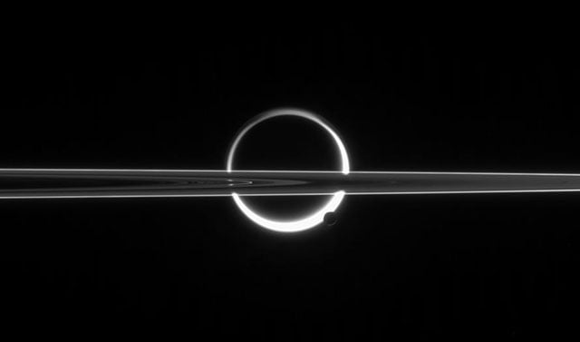 Saturn's rings cut across an eerie scene that is ruled by Titan's luminous crescent and globe-encircling haze, broken by the small moon Enceladus, whose icy jets are dimly visible at its south pole. North is up.
