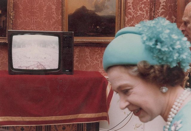Queen Elizabeth watching a small television broadcasting images of the massive crowd outside.