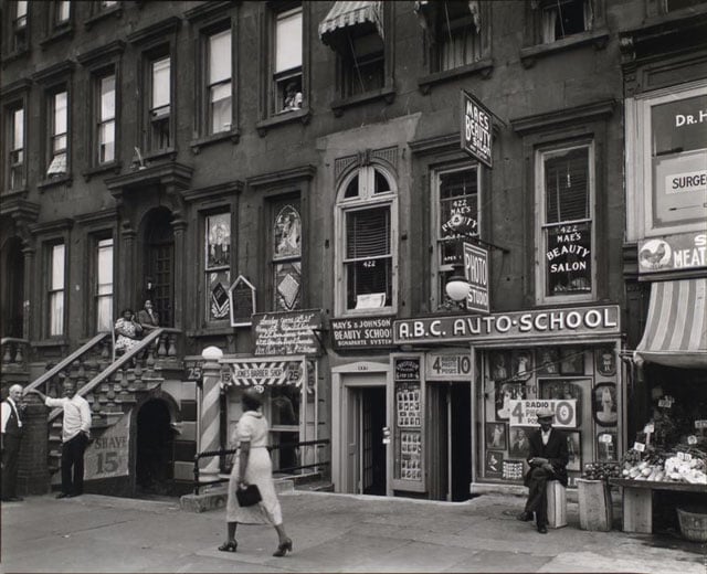 Harlem Street: II. 422-424 Lenox Avenue, Manhattan. Women sit on steps of house that serves as a church, with barber- shop below, white man talks to barber, beauty shop, and auto school next door.