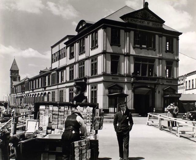 Department of Docks and Police Station, Pier A, North River, Manhattan. Long building wtih clock tower at far end housing Dept. of Docks and a Police station; man walks toward camera near newsstand in foreground.