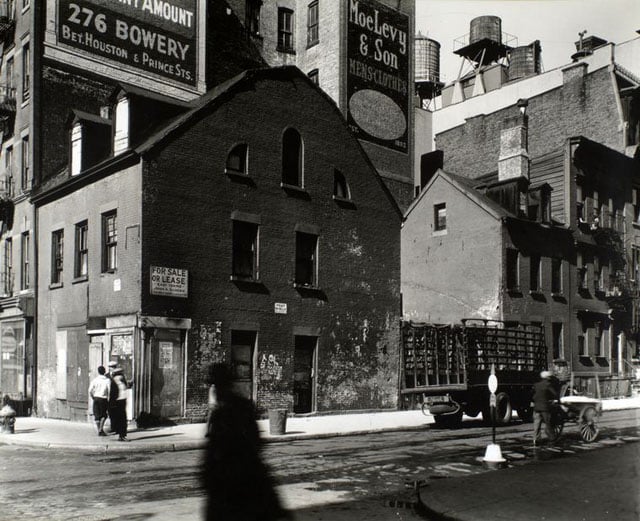 Mulberry and Prince Streets, Manhattan. Building with gambrel roof, dormers on corner, small houses along street where man pushes cart, large buildings topped with water towers beyond.