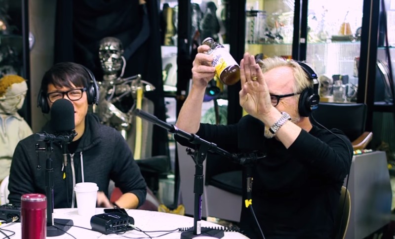 Adam Savage demonstrating how he fed the lens into a band saw, while his friend (and owner of the lens) Norman Chan cringes in horror.