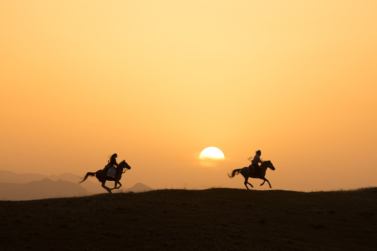 Anna and Ali riding their horse on gallop at the top of the hills as the sun sets. The Alborz mountains, a high mountain range of northern Iran can be seen in the background.