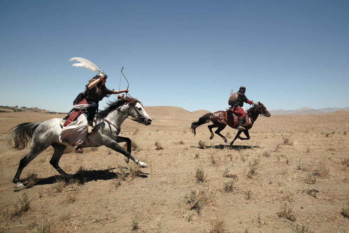 Anna shooting an arrow at Ali as she wears a kolahxud, a traditionnal Persian warrior helmet.