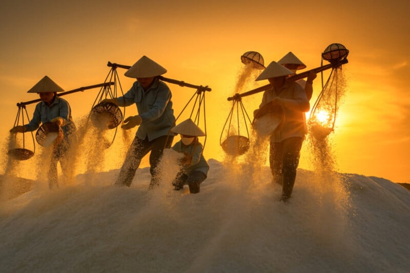 Four workers in conical hats carry baskets of salt on their shoulders, silhouetted against a golden sunset sky as salt pours from their baskets onto a mound below.