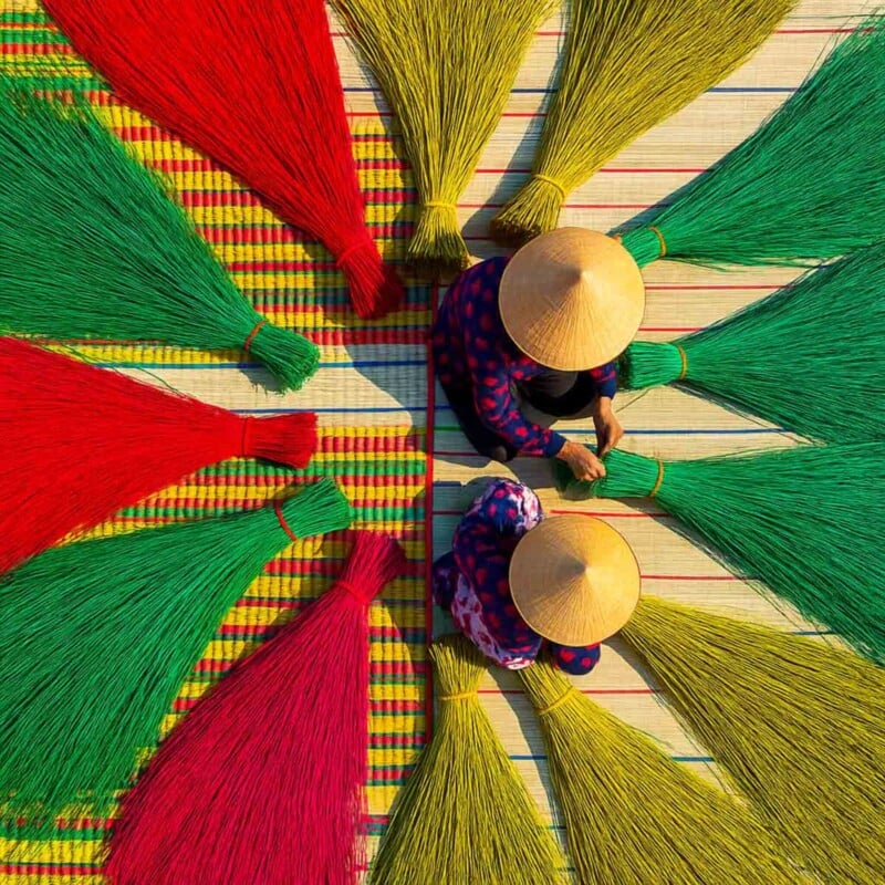 Two people wearing conical hats work among bundles of red, green, and yellow reeds arranged in a circular pattern on a woven mat, viewed from above.