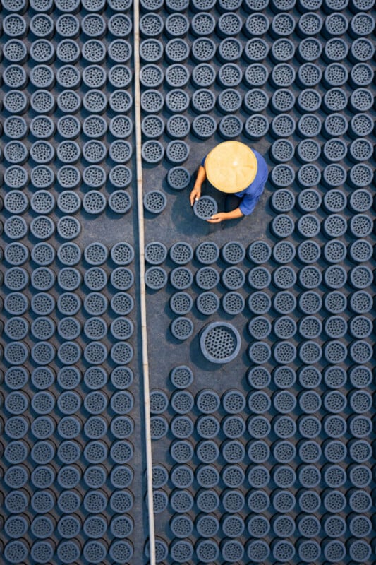 A person wearing a conical hat arranges numerous circular objects on the ground, viewed from above. The objects are arranged in a neat, grid-like pattern, with a dividing line running vertically through the image.