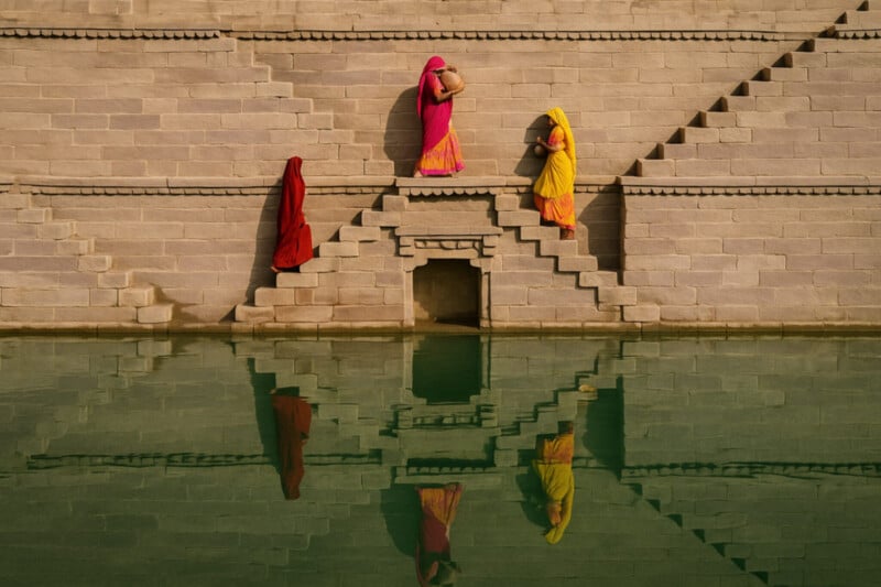 Three women in colorful traditional clothing walk along symmetrical stone steps by green water, their reflections visible in the water below. The geometric stair patterns create a striking visual effect.