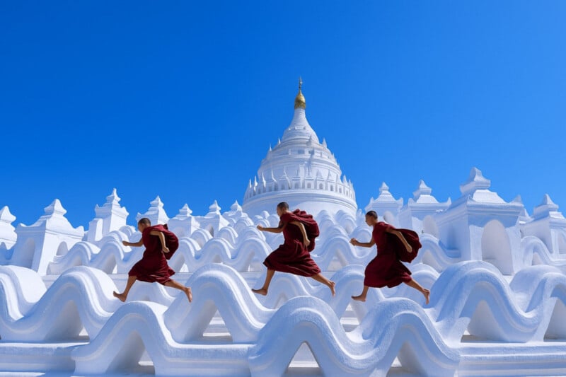 Three Buddhist monks in red robes run across the wavy white terraces of a temple with a white stupa against a clear blue sky.