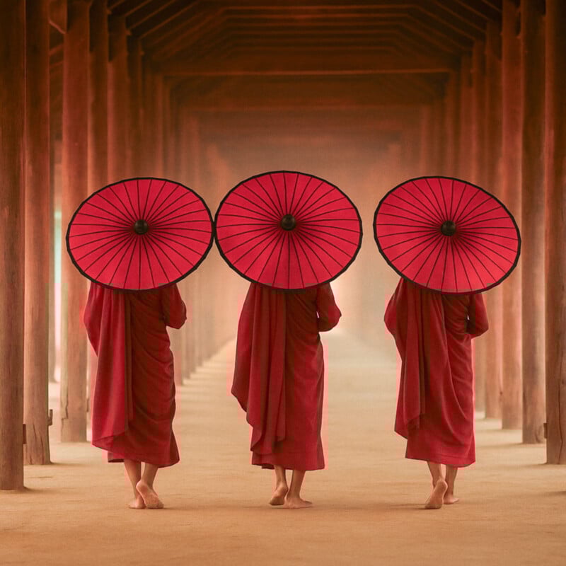 Three monks in red robes walk barefoot under wooden arches, each holding a large red umbrella that covers their heads and shoulders, creating a sense of symmetry and tranquility.