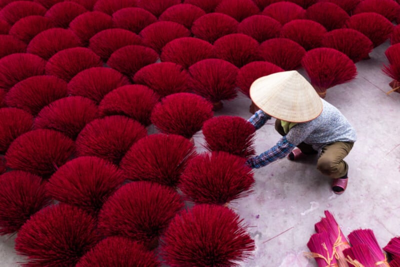 A person wearing a conical hat arranges vibrant red bundles of incense sticks, which are spread out evenly on the ground to dry. The scene is colorful and orderly, highlighting traditional craftsmanship.