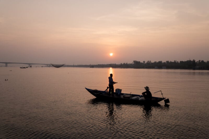 Two people are fishing on a small wooden boat at sunset, with the sun low over calm water and a hazy sky. A bridge and distant shoreline are visible in the background.